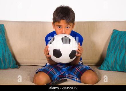 Lifestyle-Porträt zu Hause von jungen 7 oder 8 Jahre alten Jungen hält Fußball beobachten aufgeregt und nervös Fußballspiel im Fernsehen sitzen im Livin Stockfoto
