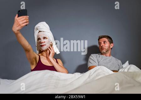 Lustige Lifestyle-Porträt von exzentrischen Hausfrau mit Make-up Gesichtsmaske und Handtuch unter Selfie im Bett und Ehemann mit verzweifelten Gesichtsausdruck in w Stockfoto