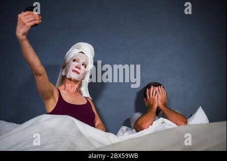 Lustige Lifestyle-Porträt von exzentrischen Hausfrau mit Make-up Gesichtsmaske und Handtuch unter Selfie im Bett und Ehemann mit verzweifelten Gesichtsausdruck in w Stockfoto