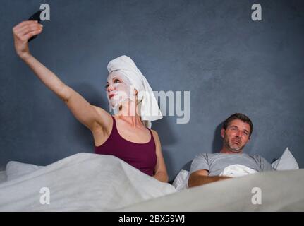 Lustige Lifestyle-Porträt von exzentrischen Hausfrau mit Make-up Gesichtsmaske und Handtuch unter Selfie im Bett und Ehemann mit verzweifelten Gesichtsausdruck in w Stockfoto