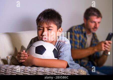 Junge traurig und gelangweilte Kind zu Hause Couch Gefühl frustriert und unbeaufsichtigt warten seinen Vater für Fußball spielen, während Mann Networking auf Handy Stockfoto