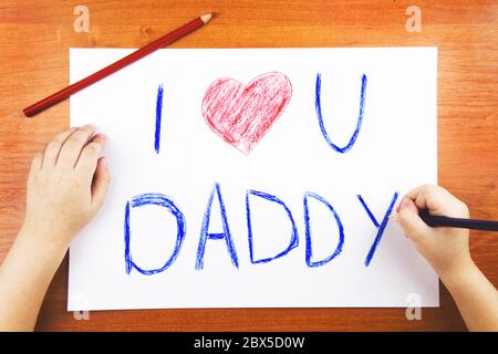 Das Konzept des Vatertags. Childs Hände Zeichnung Karte mit Worten I Love U Papa, und rotes Herz, und Farbstifte auf Holztisch. Stockfoto