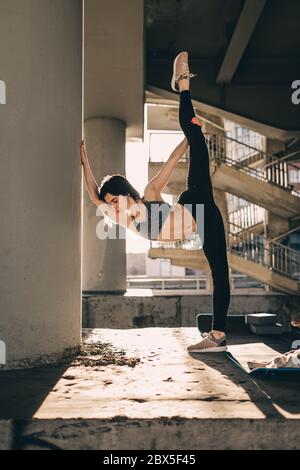 Stretching Turner oder Tänzerin Frau tun vertikale Split, Garn mit einem städtischen Blick unter der Brücke. Slim Fit Mädchen Stretching draußen auf einem Sonnenuntergang Stockfoto