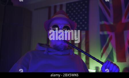 Lustige Hipster mit Sonnenbrille raucht Shisha in Neonlicht auf usa Flagge und Jack Union Hintergrund. Freizeit- und Unterhaltungskonzept Stockfoto