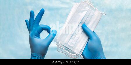 Nahaufnahme der Arzthandchen in blauen medizinischen Handschuhen, die eine chirurgische medizinische Gesichtsmaske halten und ein OK-Zeichen zeigen. Banner Panorama medizinische Konzept des Schutzes Stockfoto