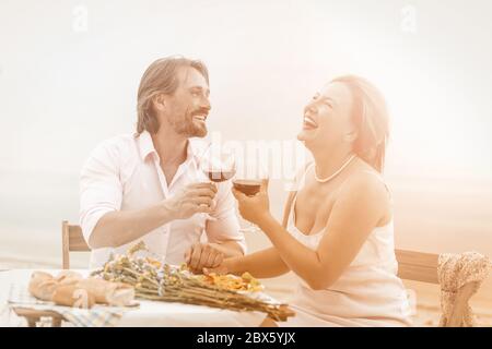 Fröhlicher Mann und Frau trinken Wein im Strandcafe. Schönes Paar lachend Hände halten und das Leben genießen, während sie zusammen im Freien sitzen. Romantische Midlife Stockfoto