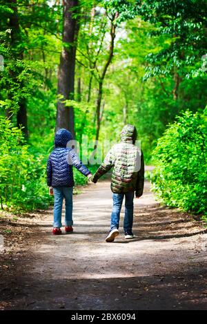 Zwei Kinder in Jacken laufen in einem sonnigen Tag in sicherer Entfernung durch den Stadtpark oder Wald und halten sich die Hände Stockfoto