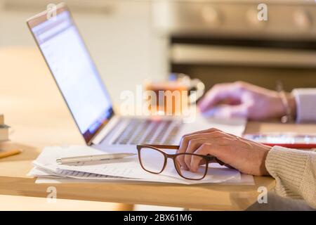 Das Bild, das den Mann über einen Laptop im Home Office sitzend und arbeitend macht, und das Klicken auf die drahtlose digitale Maus, konzentriert sich auf die Brille in der Hand. Verschwommene Tasse t Stockfoto