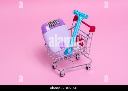 Lila Epilierer und blauer Rasierer im Supermarkt-Trolley auf einem rosa Hintergrund. Das Konzept der Wahl einer Methode der Haarentfernung. Stockfoto