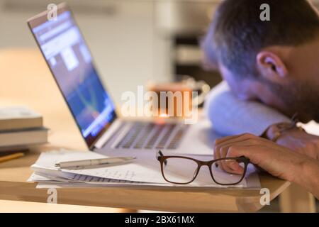 Verschwommener, müder junger kaukasischer Mann mit Brille in der Hand, einschlafen nach der Arbeit am Laptop, am Schreibtisch schlafen, auf Brille konzentrieren, Stockfoto