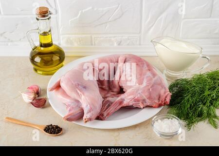 Ganze rohe Kaninchenkarkasse, saure Sahne, Knoblauch, Pfeffer, Salz, Olivenöl und Bund Dill auf Küchentisch. Zutaten für das Kochen Kaninchen Eintopf. Stockfoto