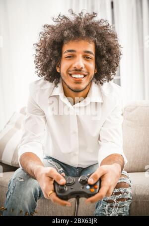 Glücklich arabischen Mann lächelt breit Computerspiel spielen. Shaggy Gamer hält Play Station Joystick sitzen auf dem bequemen Sofa zu Hause. Lustiges Zeitkonzept Stockfoto