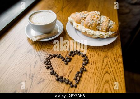 Tasse frischen Morgenkaffee und köstliche Croissant mit süßer Sahne und Herz aus Kaffeebohnen, auf Holztisch Hintergrund gemacht Stockfoto