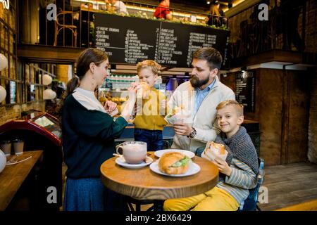 Junge fröhliche Familie, bärtigen Vater, hübsche Mutter und zwei Kinder Jungen, trinken heiße Schokolade trinken und essen traditionelle französische Croissants in einem Stockfoto