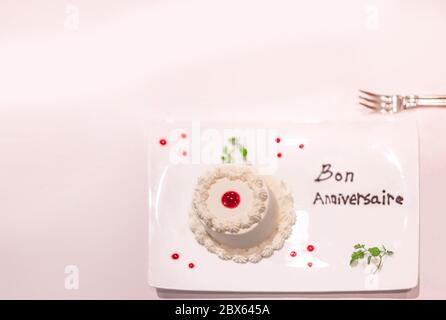 Kleiner weißer Kuchen auf einem Teller mit Erdbeersauce und Minzblättern mit Happy Birthday oder Bon Anniversaire in französisch in chocol Stockfoto