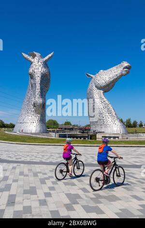 Zwei Radfahrer radeln vor den riesigen Pferdeskulpturen, die Kelpies genannt werden, in der Nähe von Falkirk, Schottland Stockfoto