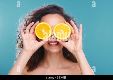 Junge glückliche schöne Frau mit dunklen lockigen Haaren, die zwei Hälften saftigen Orange an ihren Augen halten, während sie sich isoliert um das Gesicht kümmert Stockfoto