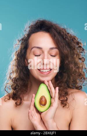 Glückliche junge luxuriöse Frau mit weißen toothy Lächeln hält die Hälfte der frischen Avocado, die ihr hilft, um die Pflege von Haut und Körper zu nehmen Stockfoto