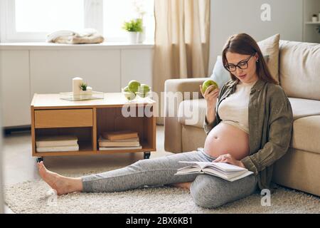 Junge schwangere erholsame Frau in Freizeitkleidung Lesen Buch und essen grünen Apfel während auf dem Boden von der Couch zu Hause sitzen Stockfoto