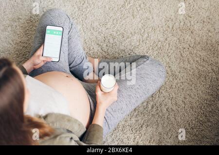 Übersicht über junge Frau mit gekreuzten Beinen, die auf einem grauen, flauschigen Teppich im Wohnzimmer sitzt, ein Glas Milch hat und im Smartphone scrollt Stockfoto