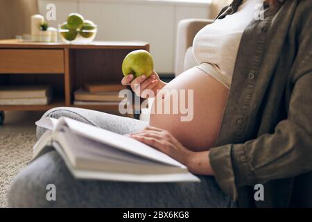 Junge entspannte schwangere Frau in Freizeitkleidung essen frischen Apfel und Buch lesen, während sie in der häuslichen Umgebung sitzen Stockfoto