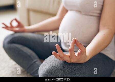 Hand und Bein im Knie der jungen schwangeren Frau in activewear sitzen in Pose des Lotus und meditieren zu Hause auf dem Boden gebogen Stockfoto