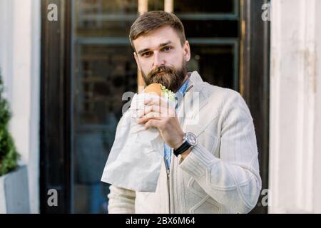 Junge schöne kaukasische bärtige Mann genießen Mittagspause, während sie frische Croissant stehen im Freien auf dem Hintergrund der Cafétür Stockfoto