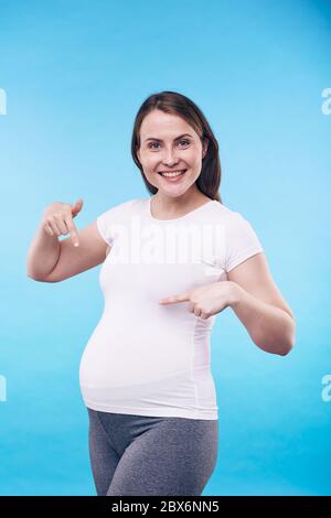 Fröhliche schwangere Frau in activewear zeigt auf ihren Bauch und zum Ausdruck Freude zu erwarten Baby vor blauem Hintergrund Stockfoto