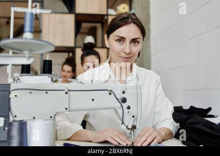 Junge zeitgenössische Näherin, die Sie anschaut, während sie am Arbeitsplatz mit einer elektrischen Nähmaschine sitzt und Kleidung anstellt oder repariert Stockfoto