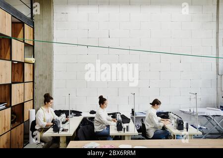 Drei junge zeitgenössische Designerinnen von Mode nähen Artikel für neue Sesonal-Kollektion während sitzen an Arbeitsplätzen in der Werkstatt Stockfoto