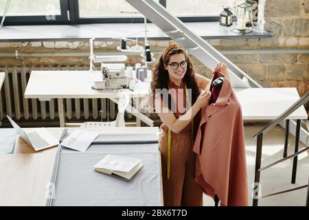 Hübsche Modedesignerin, die über neue saisonale Kollektion von Mänteln in ihrem Atelier oder in ihrer Werkstatt arbeitet, während sie von Schaufensterpuppe steht Stockfoto