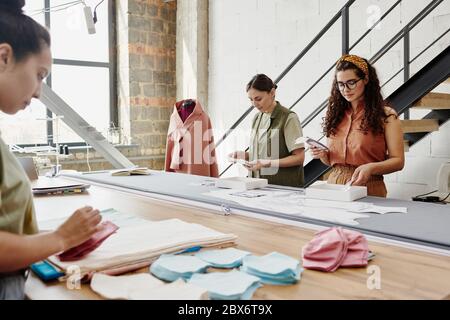 Gruppe von drei jungen Designern von Kleidung stehen an einem großen Tisch in der Werkstatt und schauen durch Proben von Stoff und andere Sachen Stockfoto