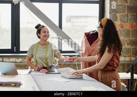 Junge fröhliche Näherin mit Maßband vorbei an ihrem Kollegen Stück rosa Tuch über den Tisch, um neue Schulterpolster machen Stockfoto