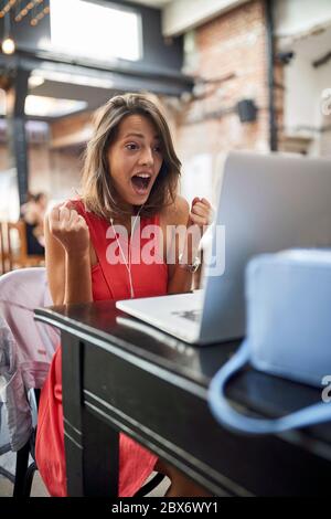 Entzückte junge Frau, die Inhalte auf ihrem Laptop mit Kopfhörern in den Ohren und Fäusten in YES Position beobachtet Stockfoto