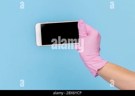 Weibliche Hand in einem rosa medizinischen Handschuh hält ein leeres modernes Handy auf einem blauen Hintergrund. Mock-up Stockfoto