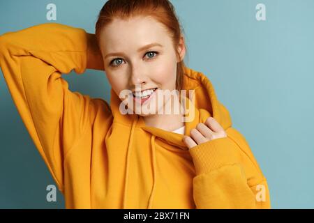 Ein Rotschopf-Mädchen in einem gelben Sweatshirt steht auf blauem Hintergrund und schaut fröhlich in den Rahmen, die Kapuze verstellend Stockfoto