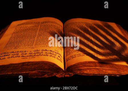 Alte hebräische Bibel im Licht brennender Kerze auf dunklem Hintergrund. Schatten aus Menora auf offenes jüdisches Gebetsbuch in der Dunkelheit. Nahaufnahme Stockfoto