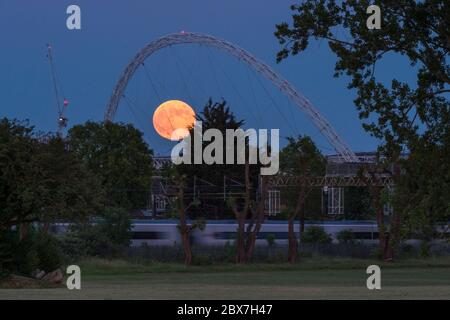 Wembley, Großbritannien. Juni 2020. UK Wetter - EIN Zug fährt vorbei, als der Vollmond im Juni, bekannt als Erdbeermond, hinter dem Wembley Stadium im Nordwesten Londons aufsteigt. Der Vollmond im Juni war laut Almanach des alten Farmers das Signal für die Indianer der Algonquin-Stämme, wilde Erdbeeren zu ernten. Der Vollmond dieses Monats fällt auch mit einer subtilen Halbfinsternis zusammen, die auftritt, wenn die Erde einen leichten Schatten über den Mond wirft. Kredit: Stephen Chung / Alamy Live News Stockfoto