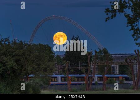 Wembley, Großbritannien. Juni 2020. UK Wetter - EIN Zug fährt vorbei, als der Vollmond im Juni, bekannt als Erdbeermond, hinter dem Wembley Stadium im Nordwesten Londons aufsteigt. Der Vollmond im Juni war laut Almanach des alten Farmers das Signal für die Indianer der Algonquin-Stämme, wilde Erdbeeren zu ernten. Der Vollmond dieses Monats fällt auch mit einer subtilen Halbfinsternis zusammen, die auftritt, wenn die Erde einen leichten Schatten über den Mond wirft. Kredit: Stephen Chung / Alamy Live News Stockfoto