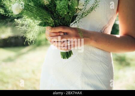 Nahaufnahme Ansicht der Frau Hände halten Brautstrauß gegen weißes Hochzeitskleid. Braut trägt Ehering. Hintergrund im Freien. Konzept für Hochzeitstag. Stockfoto