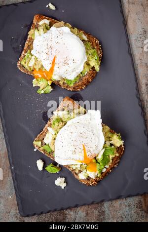 Zertrümmerte Avocado und Feta-Käse Toast mit pochierten Eiern. Blick von oben, auf dunklem Schiefer. Stockfoto