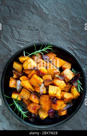 Geröstete Süßkartoffel mit roter Zwiebel und Rosmarin. Schwarze Schale, Blick von oben, über dunklem Schiefer. Stockfoto