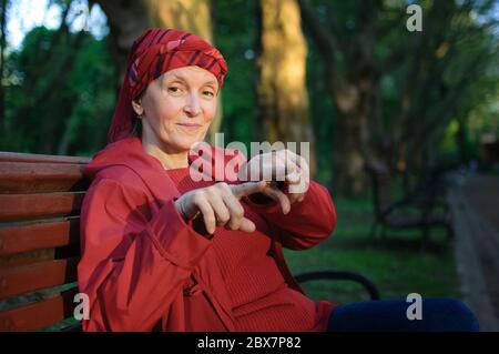 Weibliches Porträt einer reifen Frau in roter Kleidung und mit zwei Fingern auf der Bank sitzen und genießen ein gutes Wetter und Sonnenuntergang im Park Stockfoto