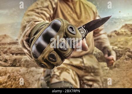 Foto eines Soldaten mit Militärausstattung, der auf dem Hintergrund des Wüstenschlachtfeldes ein kleines taktisches Messer in Handschuhen hält. Stockfoto