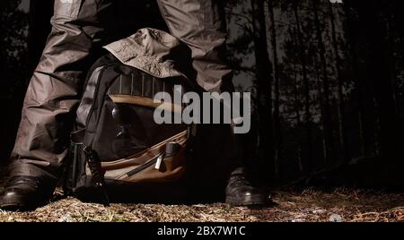 Mann in taktischem Outfit über einem Rucksack mit Camping und taktischer Ausrüstung auf Nachtwald Hintergrund stehen. Stockfoto