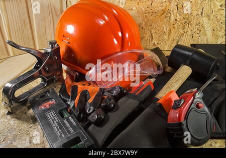 Roter Bauhelm, orange Handschuhe, Werkzeuggürtel, Regelgeräte, Hefter & Arbeitsbrille auf einem Holzbrett. Stockfoto