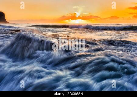 Stürmische Wellen des Pazifischen Ozeans bei Sonnenaufgang gegen die heiße orangefarbene Sonne, die über Felsen an den nördlichen Stränden von Sydney rollt. Stockfoto