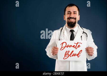 Porträt des männlichen Arztes mit Stethoskop in medizinischer Uniform, das ein weißes Papier hält und auf einem blauen isolierten Hintergrund lächelt. Stockfoto