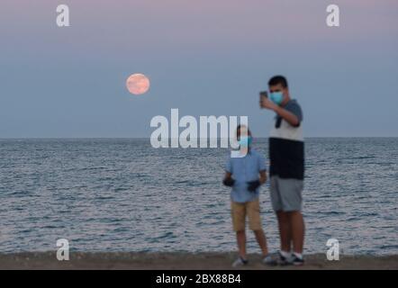 Malaga, Spanien. Juni 2020. Der Erdbeervollmond als Teil einer Mondfinsternis aus der Halbmonde erhebt sich über den Horizont, während die Menschen am Strand von Malagueta ein Selfie machen, während die Beschränkungen der Coronavirus-Krankheit (COVID-19) gelockert werden. In diesem Monat Juni sind die Erdbeer-Vollmond- und Halbmondfinsternis am 5. Juni das erste astronomische Ereignis, das die Menschen sehen können. Während dieses Monats wird auch eine Sonnenringfinsternis am 21. Juni zu sehen. Quelle: SOPA Images Limited/Alamy Live News Stockfoto