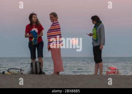 Malaga, Spanien. Juni 2020. Der Erdbeervollmond, als Teil einer Mondfinsternis, steigt über den Horizont, während die Menschen das gute Wetter am Strand von Malagueta genießen, inmitten der Lockerung der Coronavirus-Krankheit (COVID-19) Beschränkungen. In diesem Monat Juni sind die Erdbeer-Vollmond- und Halbmondfinsternis am 5. Juni das erste astronomische Ereignis, das die Menschen sehen können. Während dieses Monats wird auch eine Sonnenringfinsternis am 21. Juni zu sehen. Quelle: SOPA Images Limited/Alamy Live News Stockfoto